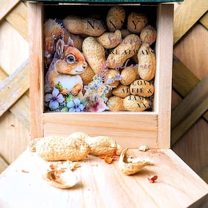 May include: A wooden squirrel feeder with a clear glass front. The feeder is filled with peanuts and has a sign that reads "Nanny's Squirrels are always welcome. Love Alfie & Jamie xxx".