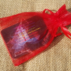 May include: A red organza bag with a drawstring closure containing a set of chakra cards. The cards feature a colorful illustration of a person meditating with the seven chakras represented by different colors.