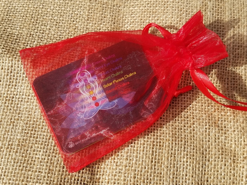 May include: A red organza bag with a drawstring closure containing a set of chakra cards. The cards feature a colorful illustration of a person meditating with the seven chakras represented by different colors.