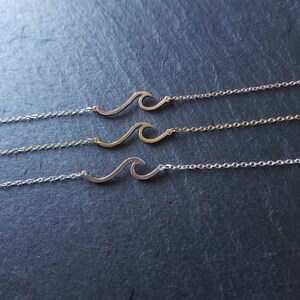 Pode incluir: Três colares delicados com pingentes em forma de onda. Os colares estão sobrepostos em uma superfície cinza escura. O colar superior tem uma corrente e um pingente de ouro rosa, o colar do meio tem uma corrente e um pingente de ouro, e o colar inferior tem uma corrente e um pingente de prata.