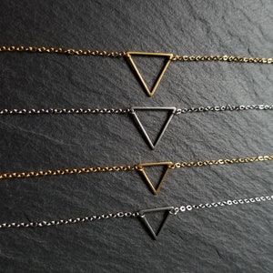 Puede incluir: Cuatro delicados collares de oro y plata con colgantes triangulares. Los collares están superpuestos sobre un fondo gris oscuro.