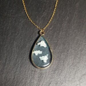 Peut inclure: Un collier en chaîne dorée avec un pendentif en forme de larme. Le pendentif est rempli de résine transparente et contient trois petits nuages blancs.