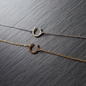 Puede incluir: Dos delicados collares con colgantes de herradura. El collar superior tiene una cadena plateada y un colgante de herradura plateada. El collar inferior tiene una cadena dorada y un colgante de herradura dorada.
