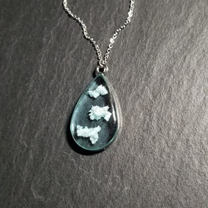 Peut inclure: Un collier en chaîne argent avec un pendentif en forme de larme. Le pendentif est rempli de résine transparente et contient de petites paillettes blanches.