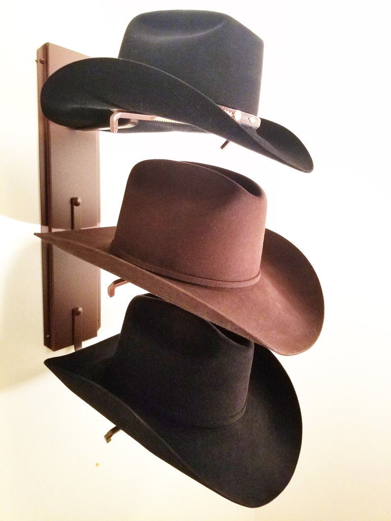 Peut inclure: Trois chapeaux de cowboy accroch&eacute;s &agrave; un support en bois. Le chapeau du haut est noir, le chapeau du milieu est marron et le chapeau du bas est noir.