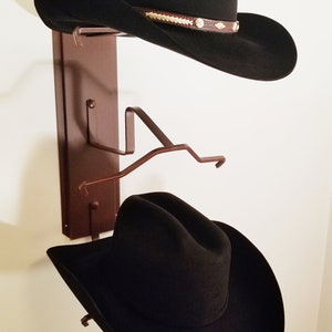 Peut inclure: Deux chapeaux de cowboy noirs accroch&eacute;s &agrave; un porte-chapeau en m&eacute;tal brun mont&eacute; sur un mur blanc.