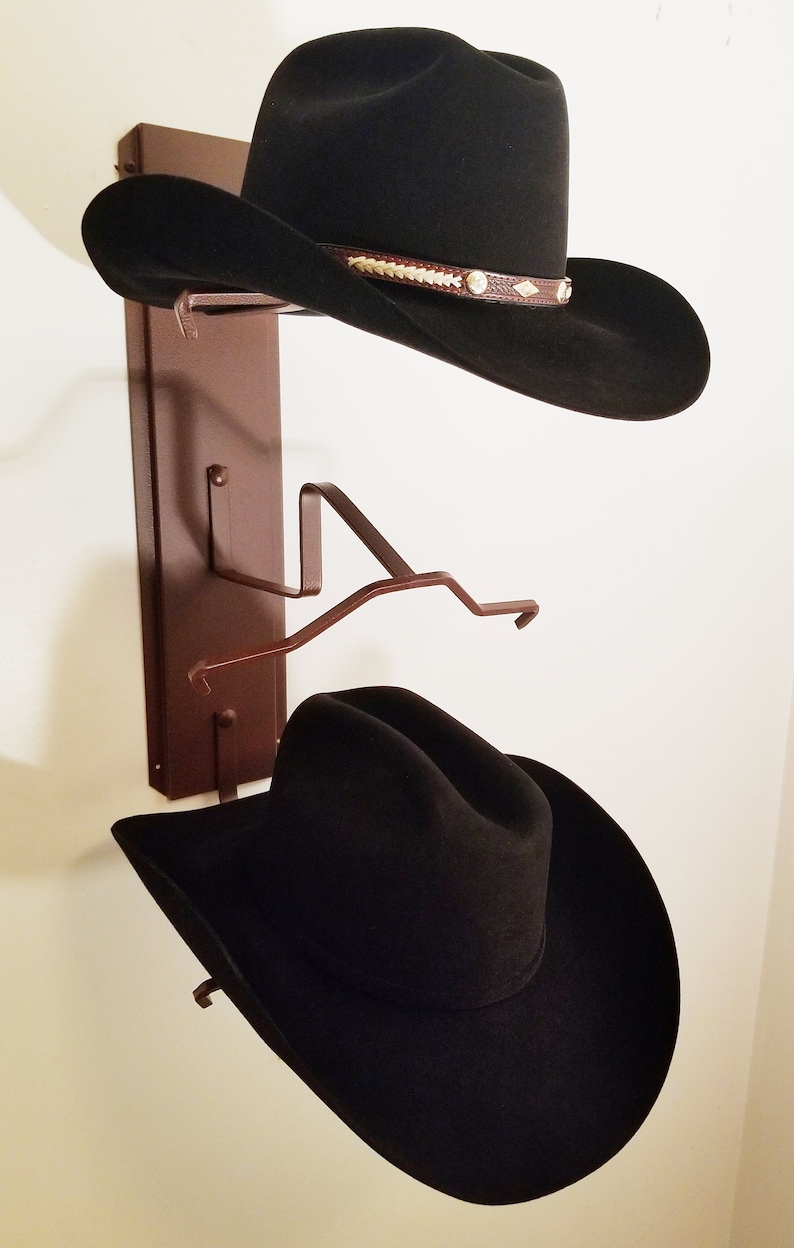 Peut inclure: Deux chapeaux de cowboy noirs accroch&eacute;s &agrave; un porte-chapeau en m&eacute;tal brun mont&eacute; sur un mur blanc.