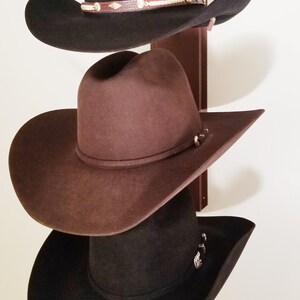 Peut inclure: Trois chapeaux de cowboy accroch&eacute;s &agrave; un support en bois. Le chapeau du haut est noir avec un bandeau en cuir marron et des accents argent&eacute;s. Le chapeau du milieu est marron et le chapeau du bas est noir.