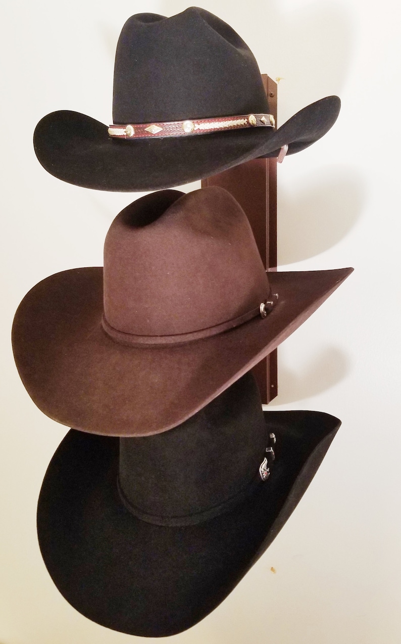 Peut inclure: Trois chapeaux de cowboy accroch&eacute;s &agrave; un support en bois. Le chapeau du haut est noir avec un bandeau en cuir marron et des accents argent&eacute;s. Le chapeau du milieu est marron et le chapeau du bas est noir.