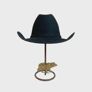 May include: A black cowboy hat with a wide brim is displayed on a decorative stand. The stand features a bronze-colored bear figurine at the base and a brown metal rod supporting the hat.