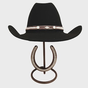May include: A black cowboy hat with a decorative band featuring white and silver accents. The hat is displayed on a horseshoe-shaped stand with a decorative border of small, silver-colored embellishments.