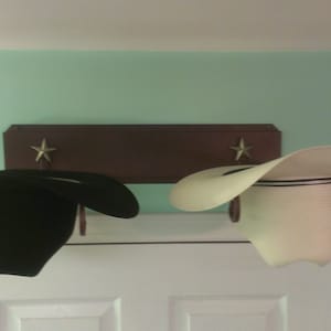 K&ouml;nnte beinhalten: Zwei Cowboyh&uuml;te, einer schwarz und einer wei&szlig;, h&auml;ngen an einem Holzregal mit Sternakzenten. Die H&uuml;te werden &uuml;ber einer wei&szlig;en T&uuml;r vor einer hellgr&uuml;nen Wand pr&auml;sentiert. Das Regal hat dekorative Metallhaken.