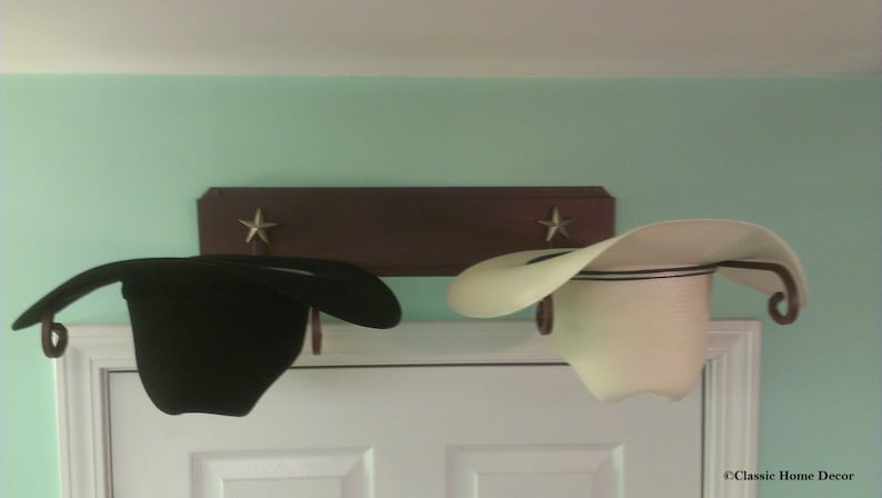 K&ouml;nnte beinhalten: Zwei Cowboyh&uuml;te, einer schwarz und einer wei&szlig;, h&auml;ngen an einem Holzregal mit Sternakzenten. Die H&uuml;te werden &uuml;ber einer wei&szlig;en T&uuml;r vor einer hellgr&uuml;nen Wand pr&auml;sentiert. Das Regal hat dekorative Metallhaken.