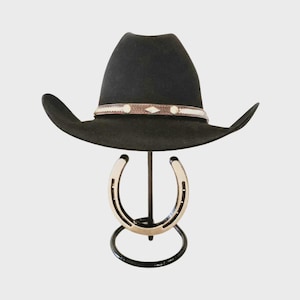 Op de afbeelding: Een zwarte cowboyhoed met een decoratieve band met witte en bruine accenten. De hoed wordt gepresenteerd op een zwarte metalen standaard met een hoefijzerontwerp. De standaard is ongeveer 30 cm hoog.