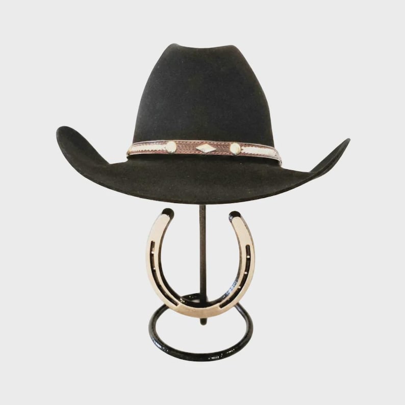 Op de afbeelding: Een zwarte cowboyhoed met een decoratieve band met witte en bruine accenten. De hoed wordt gepresenteerd op een zwarte metalen standaard met een hoefijzerontwerp. De standaard is ongeveer 30 cm hoog.