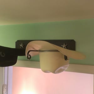 K&ouml;nnte beinhalten: Eine Wand-Hutablage mit zwei Cowboyh&uuml;ten. Ein Hut ist schwarz, der andere cremefarben. Die Ablage ist dunkelbraun mit Sternaussparungen. Die H&uuml;te werden &uuml;ber einem wei&szlig;en T&uuml;rrahmen ausgestellt.