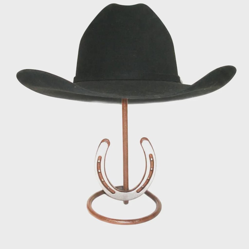May include: A black cowboy hat displayed on a horseshoe-shaped hat stand. The hat has a wide brim and a rounded crown. The stand is made of metal with a horseshoe design in the center.
