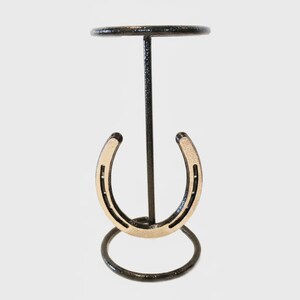 Op de afbeelding: Een zwarte metalen standaard met een goudkleurige hoefijzer. De standaard heeft een cirkelvormige basis en een kleine, ronde bovenkant, ontworpen om een klein object vast te houden. De standaard is van metaal.