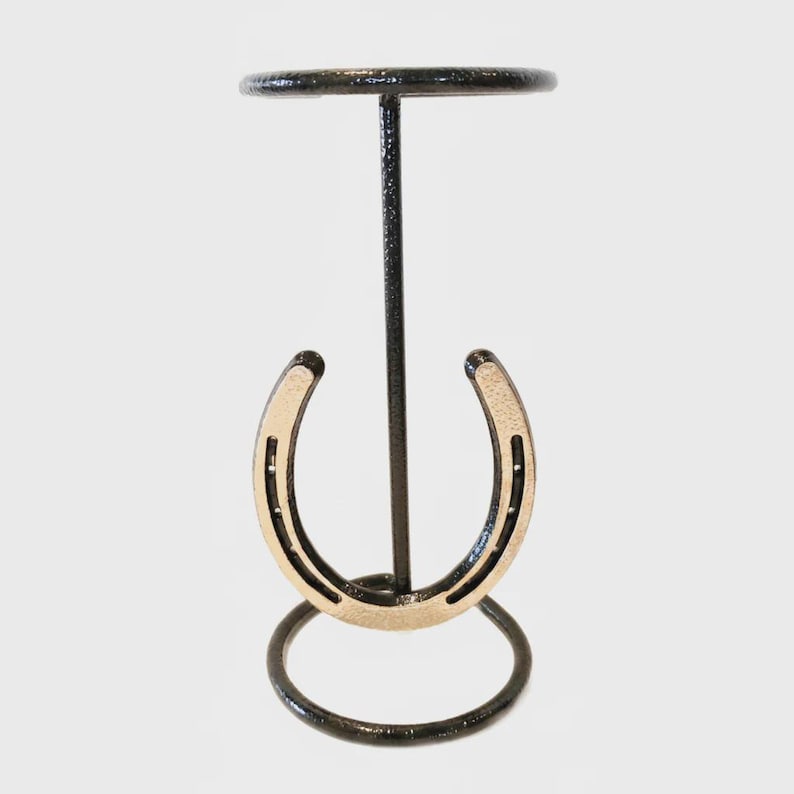Op de afbeelding: Een zwarte metalen standaard met een goudkleurige hoefijzer. De standaard heeft een cirkelvormige basis en een kleine, ronde bovenkant, ontworpen om een klein object vast te houden. De standaard is van metaal.
