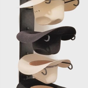 May include: A black metal hat rack displaying five cowboy hats. The hats are in various colors, including brown, black, and cream. Each hat is held by a curved metal hook. The rack is mounted on a white wall.