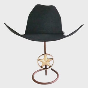 May include: A black cowboy hat displayed on a decorative metal stand. The stand features a bronze-colored base and a gold star emblem. The hat has a wide brim and a classic western design.