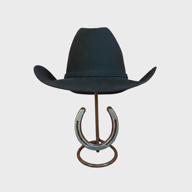 May include: A black cowboy hat with a curved brim sits atop a metal horseshoe-shaped hat stand. The hat stand has a brown metal base and a silver horseshoe design. The hat is made of a felt-like material.