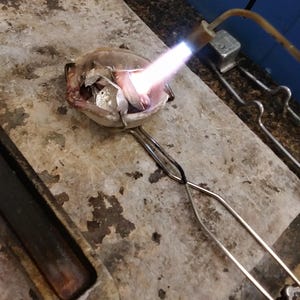May include: A small ceramic crucible containing metallic scraps is being heated by a bright blue flame from a handheld torch. The crucible is held by metal tongs over a textured, heat-resistant stone surface used for jewelry metal melting.