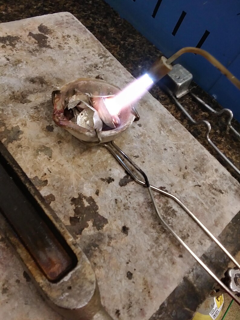May include: A small ceramic crucible containing metallic scraps is being heated by a bright blue flame from a handheld torch. The crucible is held by metal tongs over a textured, heat-resistant stone surface used for jewelry metal melting.