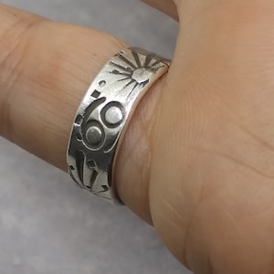 May include: A silver-toned metal ring featuring an engraved design of a sun with radiating rays and a circular symbol resembling the number sixty-nine. The band has a smooth, polished finish with dark, recessed lines highlighting the etched patterns.