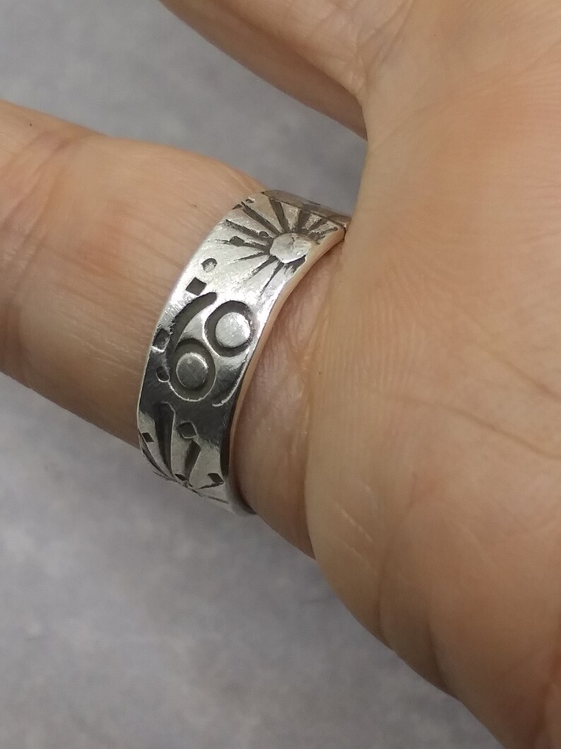 May include: A silver-toned metal ring featuring an engraved design of a sun with radiating rays and a circular symbol resembling the number sixty-nine. The band has a smooth, polished finish with dark, recessed lines highlighting the etched patterns.