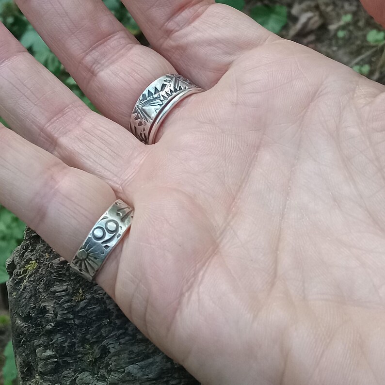 May include: Two silver rings worn on fingers. The ring on the middle finger features a geometric triangular pattern. The ring on the ring finger displays a sunburst motif and a circular symbol resembling the Cancer zodiac sign.