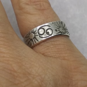 May include: A silver-toned metal ring featuring an engraved Cancer zodiac symbol in the center, flanked by two sunburst designs with radiating lines. The band has a smooth, polished finish with dark, recessed etchings for the symbols.