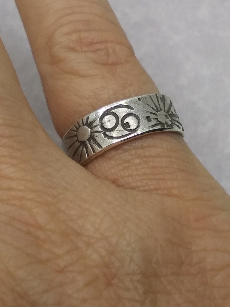 May include: A silver-toned metal ring featuring an engraved Cancer zodiac symbol in the center, flanked by two sunburst designs with radiating lines. The band has a smooth, polished finish with dark, recessed etchings for the symbols.