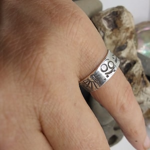 May include: A silver-toned band ring featuring a stamped design of a sunburst with rays and a circular symbol with two dots. The metal surface has a polished finish with dark recessed areas highlighting the engraved patterns.