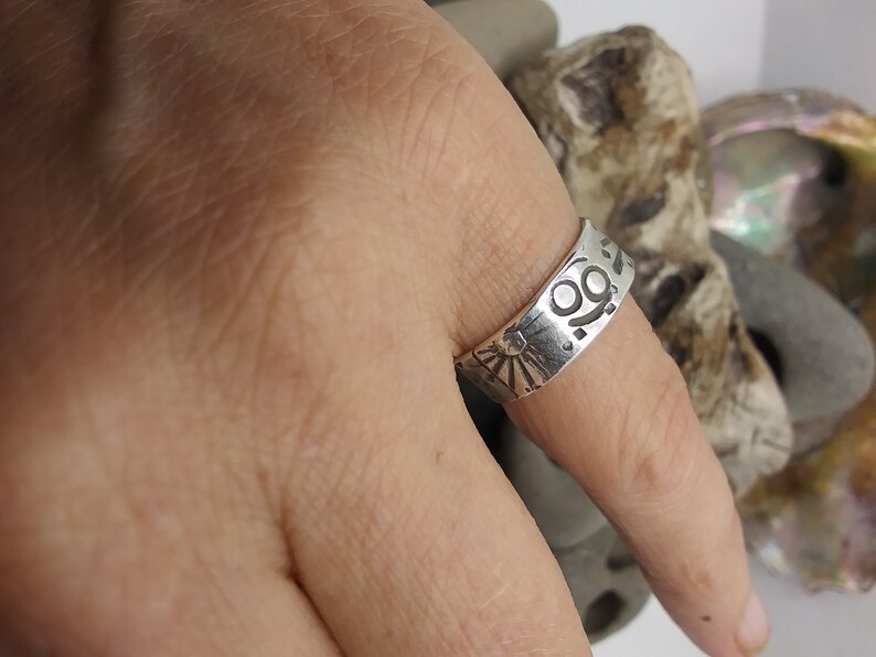 May include: A silver-toned band ring featuring a stamped design of a sunburst with rays and a circular symbol with two dots. The metal surface has a polished finish with dark recessed areas highlighting the engraved patterns.
