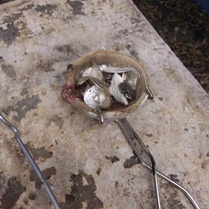 May include: A ceramic melting crucible containing various small pieces of scrap silver metal, held by a pair of long metal tongs. The crucible has a worn, off-white exterior with dark residue and a small pouring spout on the left side.