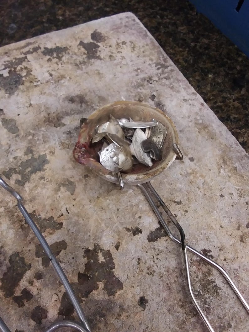 May include: A ceramic melting crucible containing various small pieces of scrap silver metal, held by a pair of long metal tongs. The crucible has a worn, off-white exterior with dark residue and a small pouring spout on the left side.