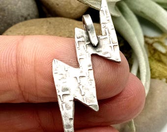 May include: A silver-colored pendant shaped like a lightning bolt with a textured, matte surface featuring small, recessed cross-shaped indentations throughout. The pendant includes a small, integrated metal loop at the top for attachment.
