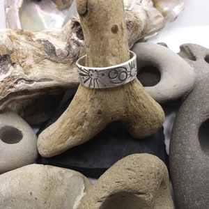 May include: A silver band ring featuring an engraved Cancer zodiac symbol and sun motifs. The ring is displayed on a piece of driftwood surrounded by various smooth, grey, and tan stones of different shapes and sizes.