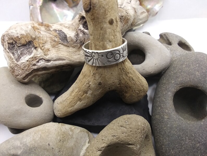 May include: A silver band ring featuring an engraved Cancer zodiac symbol and sun motifs. The ring is displayed on a piece of driftwood surrounded by various smooth, grey, and tan stones of different shapes and sizes.