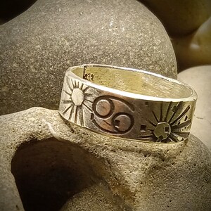 May include: A silver-toned metal ring featuring an engraved Cancer zodiac symbol flanked by two sun motifs. The band has a flat profile with dark, recessed lines highlighting the celestial designs and a visible hallmark stamp on the inner surface.