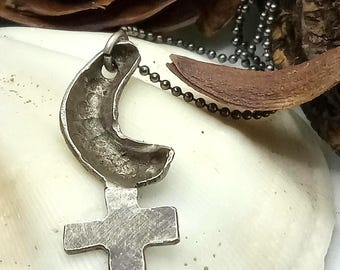 May include: A silver-toned metal pendant shaped like a crescent moon atop a cross, suspended from a thin, dark metallic ball chain. The pendant has a textured, matte finish and a visible loop at the top for the chain to pass through.