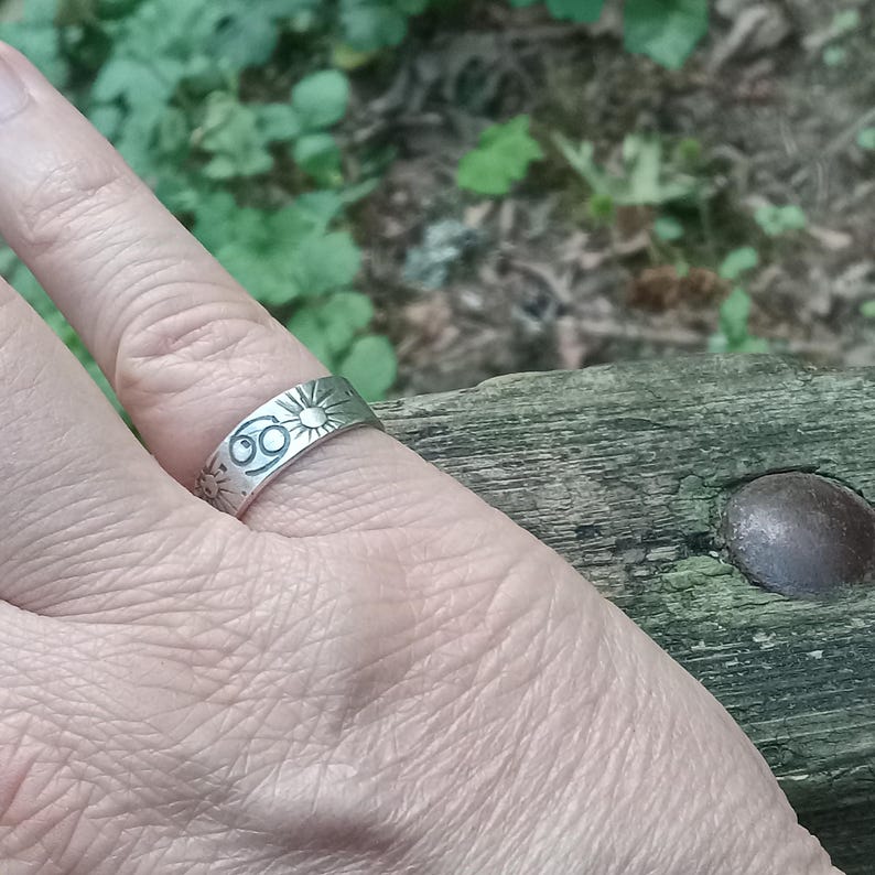 May include: A silver-toned metal ring featuring an engraved Cancer zodiac symbol and sun motifs. The band has a flat, polished surface with dark, recessed lines highlighting the celestial designs etched into the metal exterior.