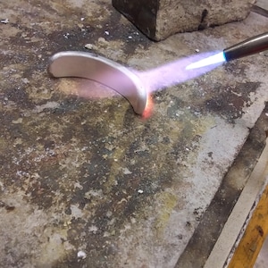 May include: A curved silver-colored metal piece being heated by a blue flame from a torch. The metal glows with a warm orange hue at the point of contact with the flame, while the rest of the surface remains a matte silver tone.