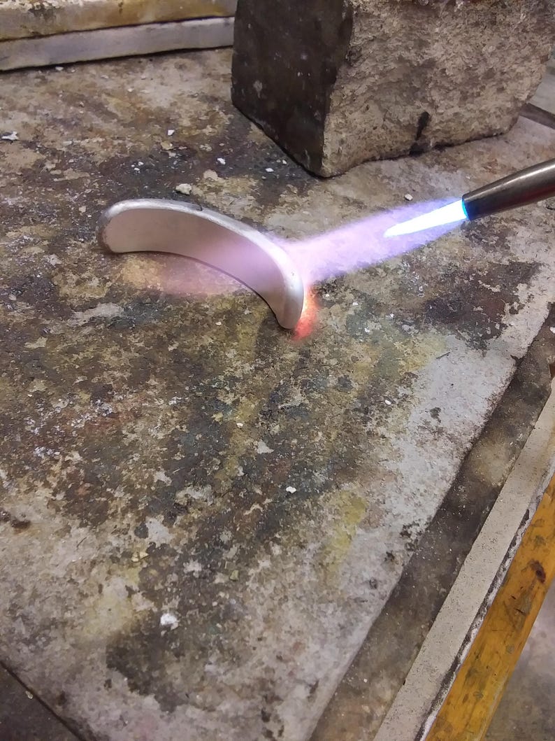 May include: A curved silver-colored metal piece being heated by a blue flame from a torch. The metal glows with a warm orange hue at the point of contact with the flame, while the rest of the surface remains a matte silver tone.