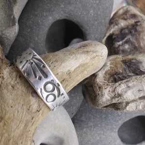 May include: A silver band ring featuring an engraved sunburst design and the Cancer zodiac symbol. The metal surface has a polished finish with dark, recessed lines highlighting the etched celestial and astrological motifs.