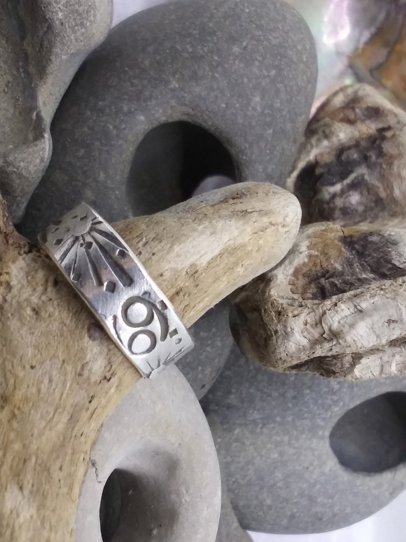 May include: A silver band ring featuring an engraved sunburst design and the Cancer zodiac symbol. The metal surface has a polished finish with dark, recessed lines highlighting the etched celestial and astrological motifs.