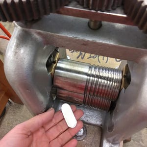 May include: A metal rolling mill machine featuring two large, stacked steel rollers with smooth and grooved sections, set within a heavy gray cast iron frame with top-mounted gears, used for flattening or shaping small, oval-shaped metal blanks.
