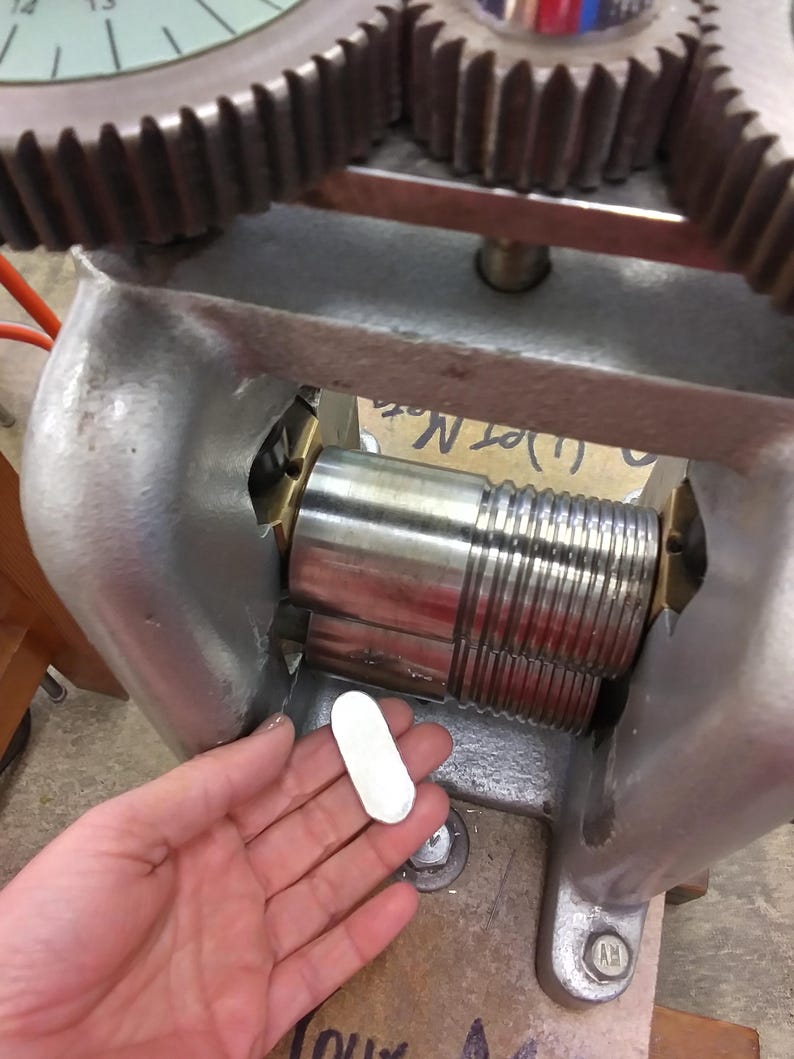 May include: A metal rolling mill machine featuring two large, stacked steel rollers with smooth and grooved sections, set within a heavy gray cast iron frame with top-mounted gears, used for flattening or shaping small, oval-shaped metal blanks.