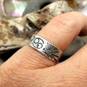 May include: A silver-toned metal ring featuring a flat band design with engraved details. The surface displays a central Cancer zodiac symbol flanked by two sunburst motifs, all rendered in a dark, oxidized finish against the silver metal.